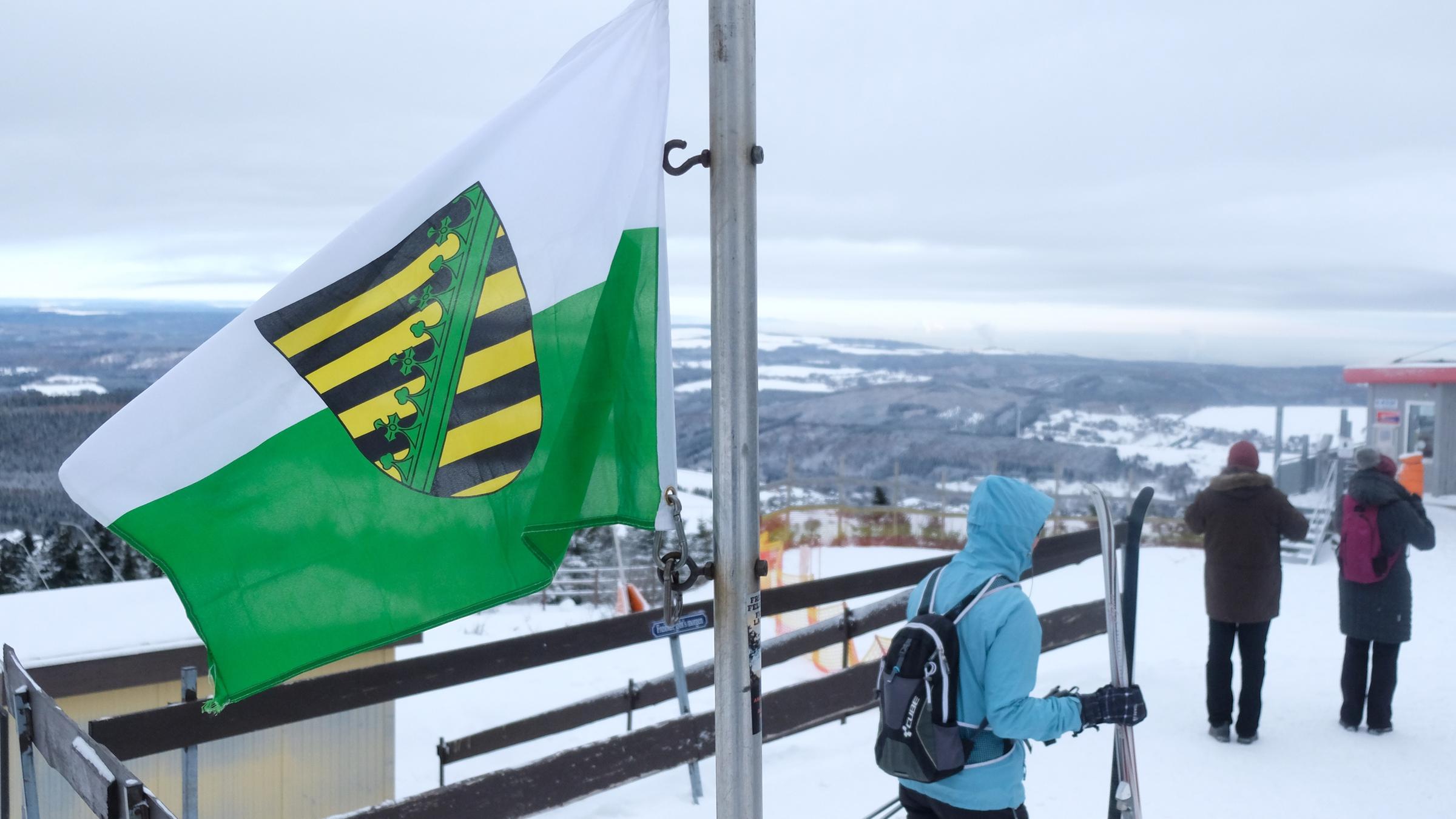 Spaziergänger und Wintersportler stehen auf dem Fichtelberg hinter einer sächsischen Landesflagge.