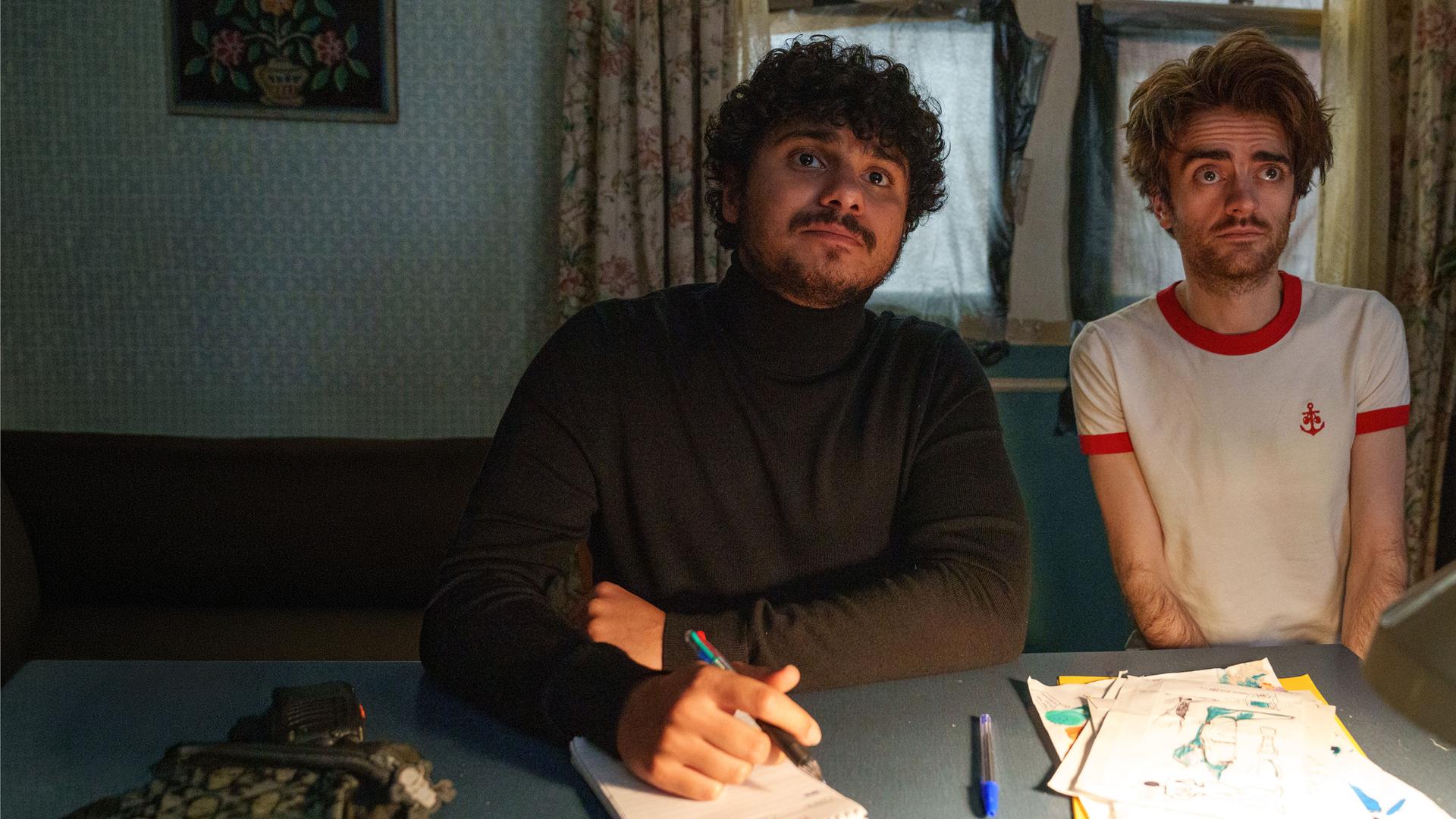 "Extraordinary - Sackgasse": Kash (Bilal Hasna) und Jizzlord (Luke Rollason) sitzen in einem Arbeitszimmer an einem Schreibtisch. Kash schaut aufmerksam geradeaus und hält dabei einen Stift in der Hand, der auf einem Block aufliegt, während Jizzlord skeptisch blickt.