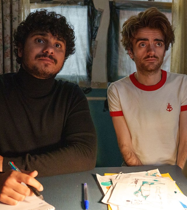 "Extraordinary - Sackgasse": Kash (Bilal Hasna) und Jizzlord (Luke Rollason) sitzen in einem Arbeitszimmer an einem Schreibtisch. Kash schaut aufmerksam geradeaus und hält dabei einen Stift in der Hand, der auf einem Block aufliegt, während Jizzlord skeptisch blickt.