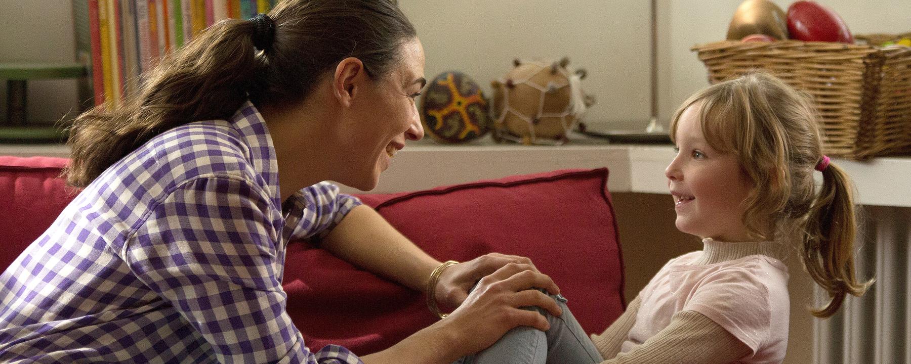 Katinka (Judith Bohle) und Ronja (Lotte Shirin Keiling) sitzen nebeneinander auf einem Sofa und reden. Katinka blickt zu Ronja und stützt ihre Hände auf Ronjas Knie. Im Hintergrund sind Bücher, sowie ein Korb mit Musikinstrumenten zu sehen.