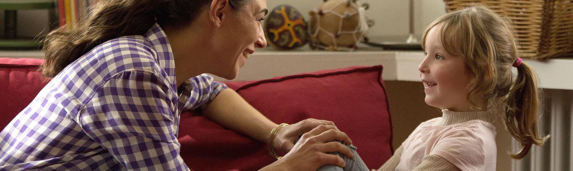 Katinka (Judith Bohle) und Ronja (Lotte Shirin Keiling) sitzen nebeneinander auf einem Sofa und reden. Katinka blickt zu Ronja und stützt ihre Hände auf Ronjas Knie. Im Hintergrund sind Bücher, sowie ein Korb mit Musikinstrumenten zu sehen.