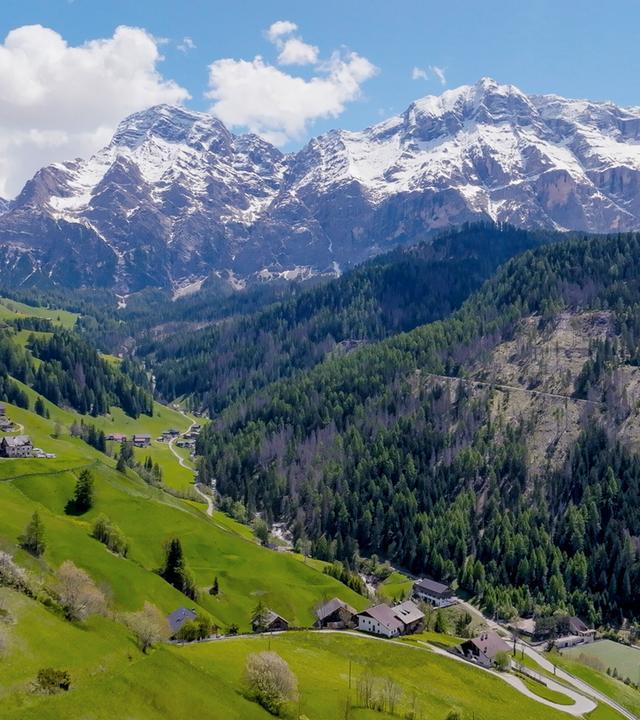 Das Bild zeigt eine atemberaubende Landschaft in Südtirol. Im Vordergrund steht eine kleine, weiße Kirche mit einem spitzen Turm, die auf einem sanften Hügel thront. Die Uhr am Turm ist deutlich sichtbar. Rund um die Kirche erstreckt sich eine weitläufige, grüne Wiese mit vereinzelt stehenden Bäumen.  Im Hintergrund erheben sich beeindruckende, schneebedeckte Berge, deren Gipfel schroff und zerklüftet sind. Diese Berge sind von einem sattgrünen Wald umgeben, der sich bis zu den Bergflanken zieht. Der Himmel ist überwiegend blau mit einigen weißen Wolken, die die sonnige Atmosphäre unterstreichen.  Auf den sanften Hügeln sind einige kleine Siedlungen zu erkennen, die aus traditionellen, mehrgeschossigen Gebäuden bestehen. Die Straßen sind geschwungen und ziehen sich durch die Landschaft. Insgesamt vermittelt das Bild eine starke Verbindung zwischen Natur und menschlicher Siedlung in dieser malerischen Region.
