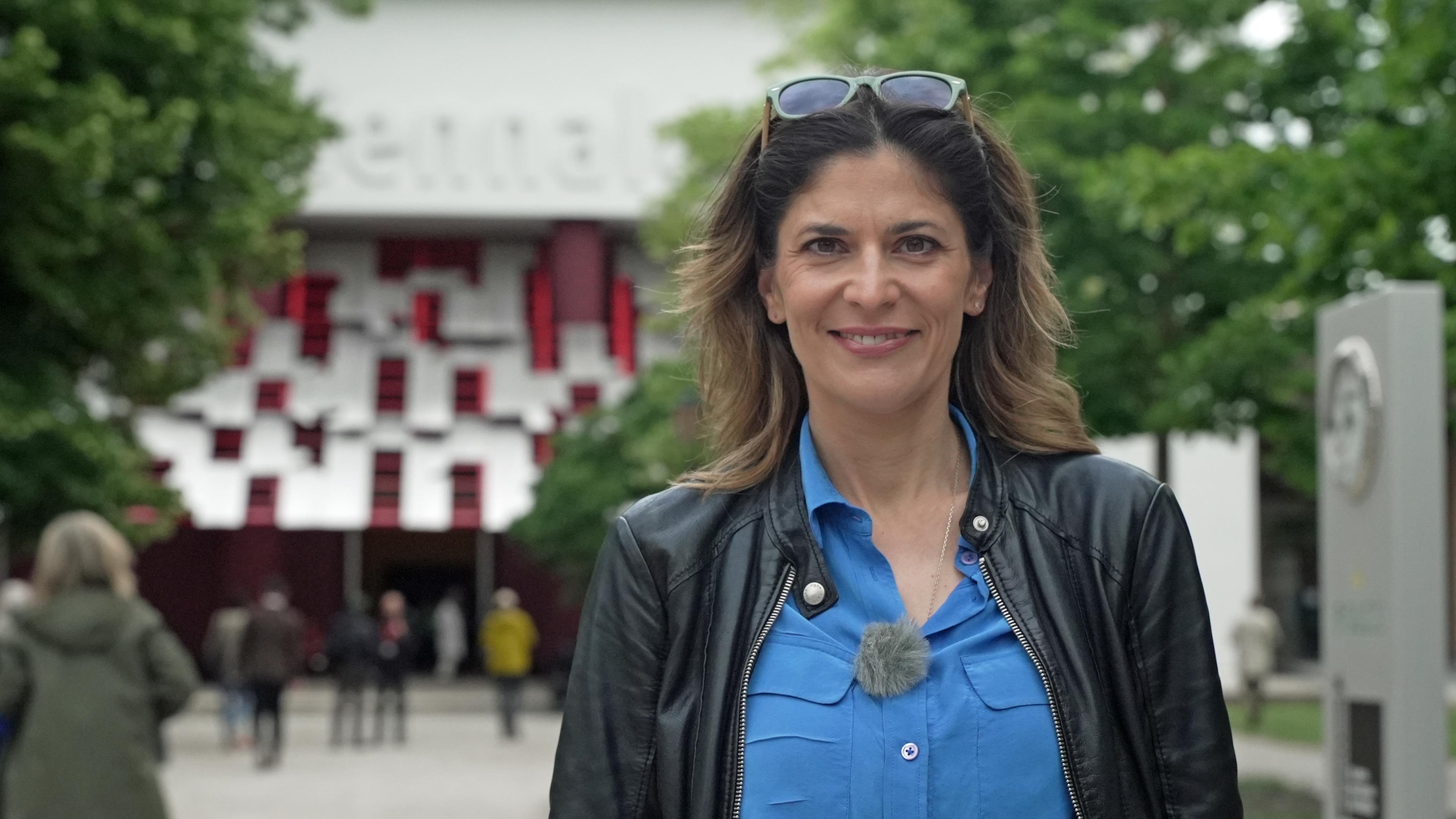 Moderatorin Katty Salié  steht vor der Hauptausstellungshalle in den Giardini und lächelt in die Kamera.