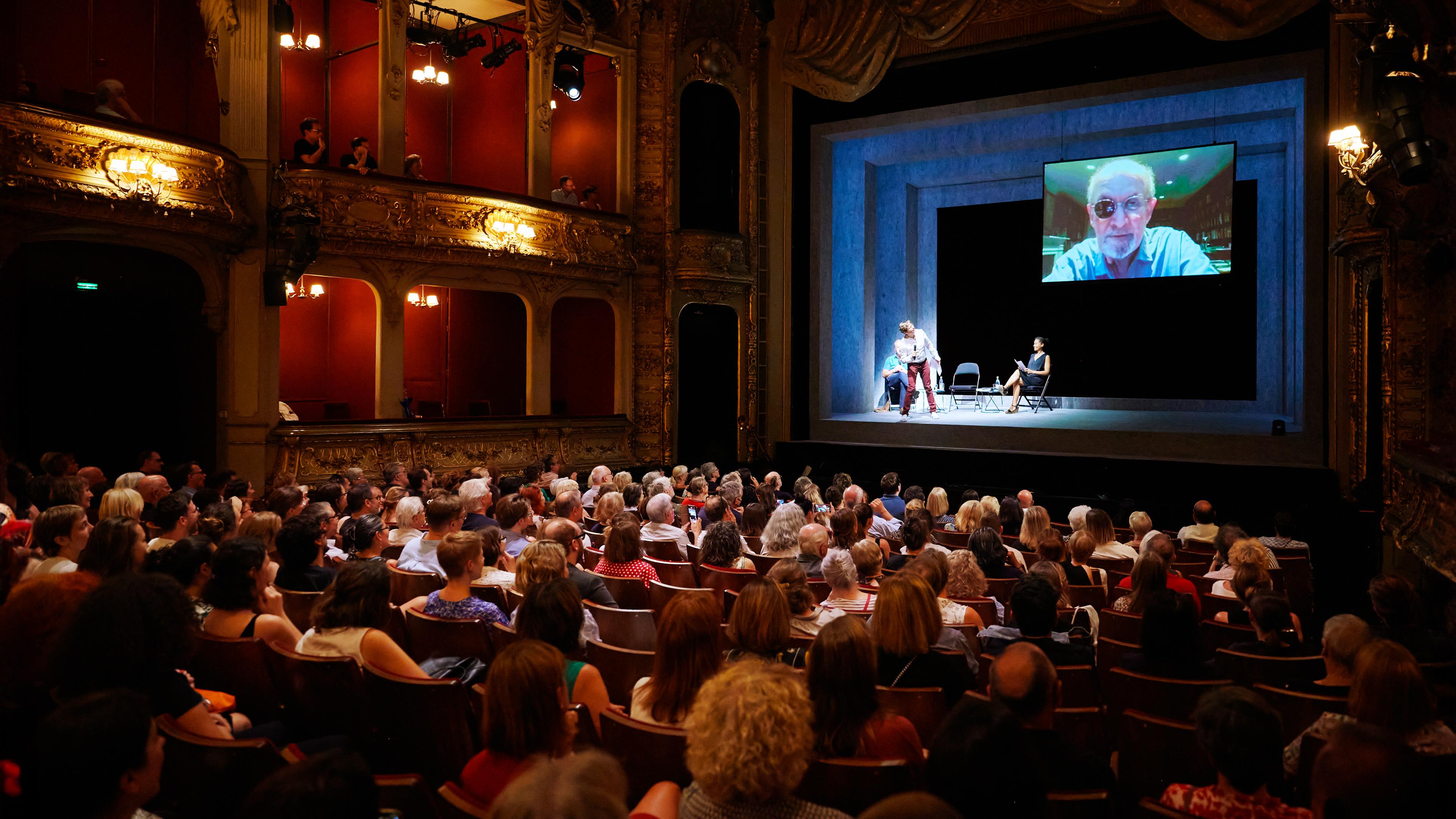 In einem großen Theatersaal sitzt Publik vor einer Bühne, auf der drei Personen sitzen und stehen. Über der Bühne hängt ein großer Monitor, auf dem ein älterer Mann mit Augenklappe zu sehen ist.