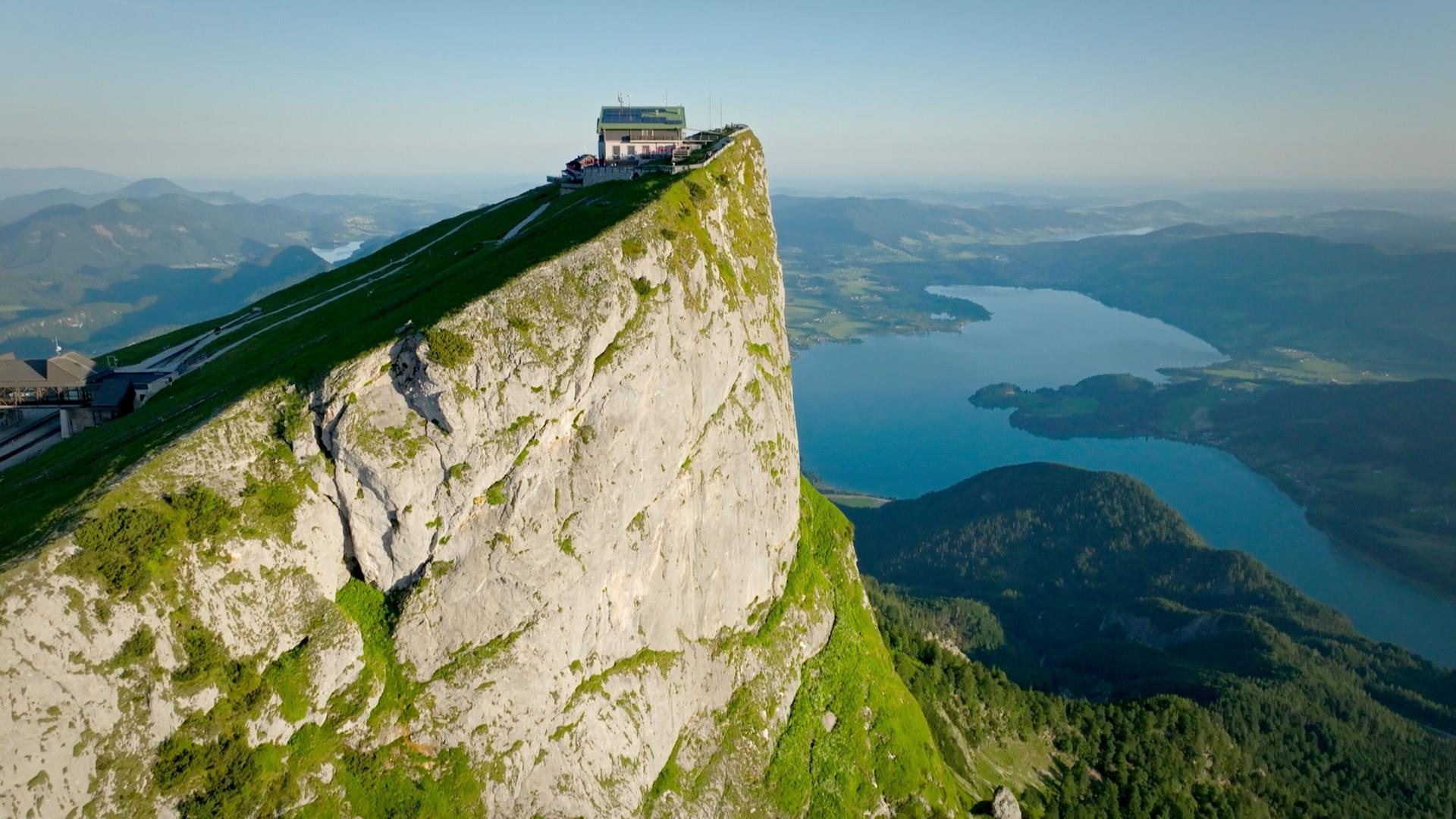 Das Bild zeigt eine beeindruckende Landschaft im Salzburger Seenland, Österreich. Im Vordergrund ist eine steile, grüne Berghang zu sehen, der zu einer markanten Felskante führt. Auf der Felskante steht ein modernes Gebäude, das vermutlich ein Hotel oder eine Aussichtsplattform ist, umgeben von einer Terrasse mit Stühlen und Tischen.  Darunter erstreckt sich ein klarer, blauer See, der von sanften Hügeln umgeben ist. Im Hintergrund sind weitere Berge sichtbar, die die Landschaft ergänzen. Die Atmosphäre wirkt ruhig und einladend, ideal für Erholungssuchende und Naturliebhaber. Die Szene vermittelt einen Eindruck von Weite und Naturschönheit, typisch für die Region Salzburg.