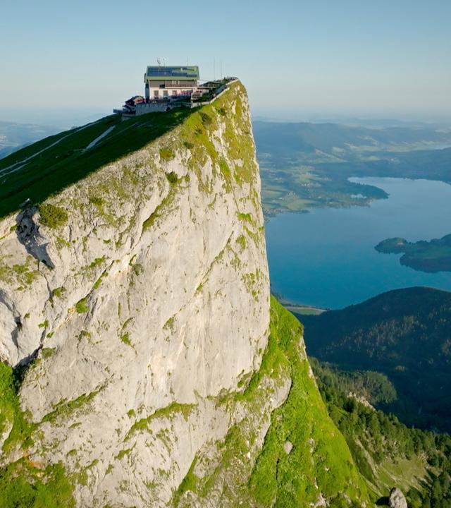 Das Bild zeigt eine beeindruckende Landschaft im Salzburger Seenland, Österreich. Im Vordergrund ist eine steile, grüne Berghang zu sehen, der zu einer markanten Felskante führt. Auf der Felskante steht ein modernes Gebäude, das vermutlich ein Hotel oder eine Aussichtsplattform ist, umgeben von einer Terrasse mit Stühlen und Tischen.  Darunter erstreckt sich ein klarer, blauer See, der von sanften Hügeln umgeben ist. Im Hintergrund sind weitere Berge sichtbar, die die Landschaft ergänzen. Die Atmosphäre wirkt ruhig und einladend, ideal für Erholungssuchende und Naturliebhaber. Die Szene vermittelt einen Eindruck von Weite und Naturschönheit, typisch für die Region Salzburg.