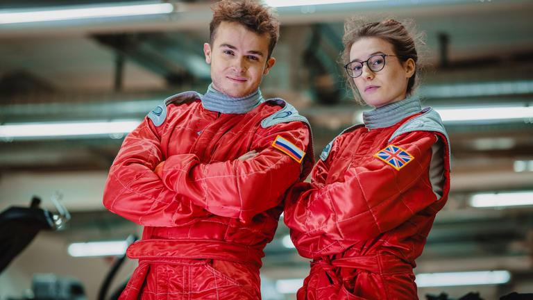 Szene aus "Umlaufbahnen" von Samantha Harvey: Ein junger Astronaut und eine junge Astronautin stehen mit verschränkten Armen nebeneinander. Sie blicken in die Kamera, die Frau trägt eine Brille. Sie sind mit roten Overalls bekleidet. Auf dem rechten Arm des Mannes ist eine russische Flagge zu sehen. Die großbritannische Flagge befindet sich auf dem rechten Arm der Frau. Der Hintergrund ist unscharf, grau und es sind mehrere Leuchtstoffröhren zu erkennen.
