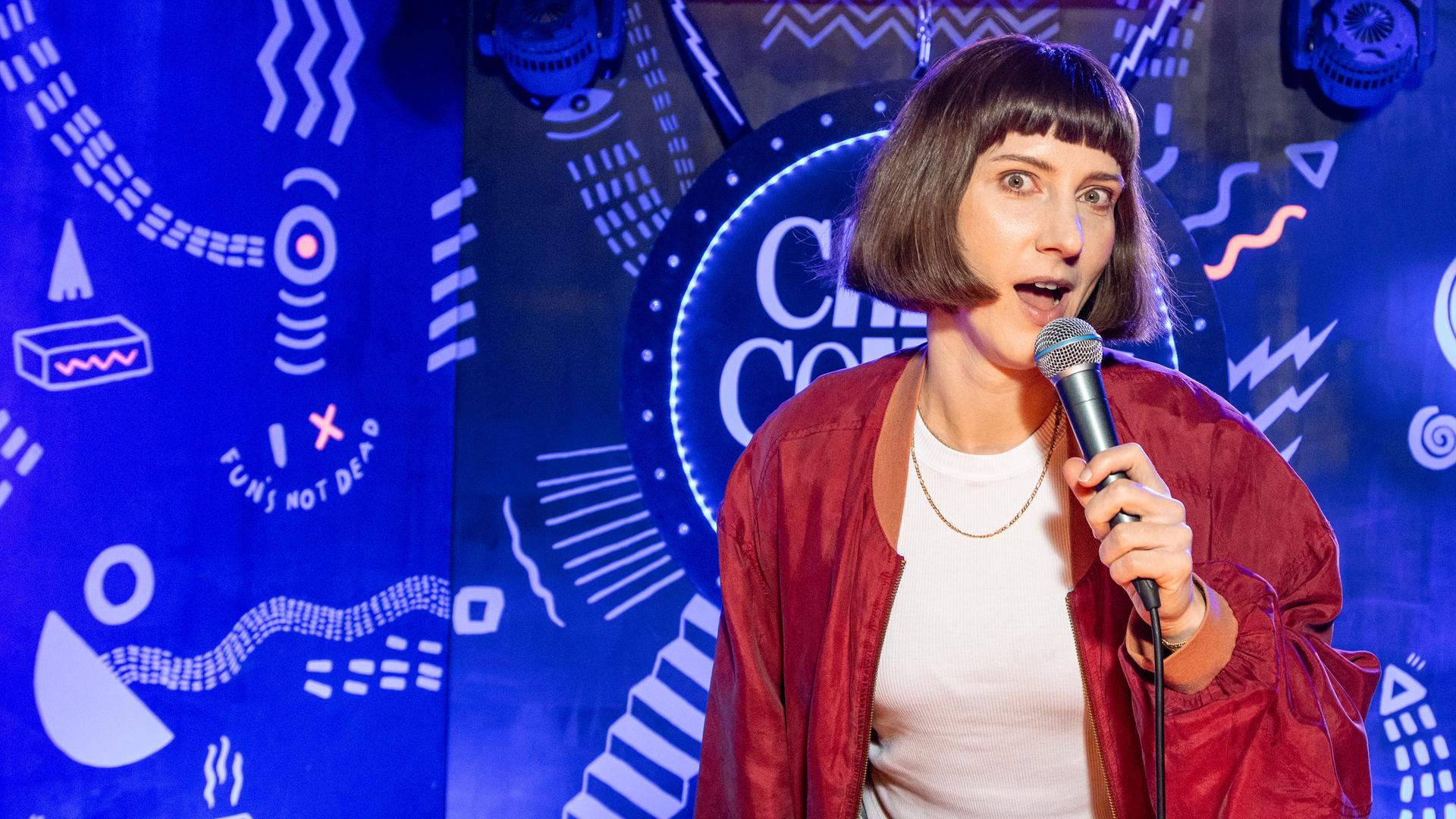 Sandra Schmidt steht auf der Mitte der Bühne und hält ein Mikrofon in der Hand. Ihr Mund ist geöffnet und der Hintergrund ist blau beleuchtet. 