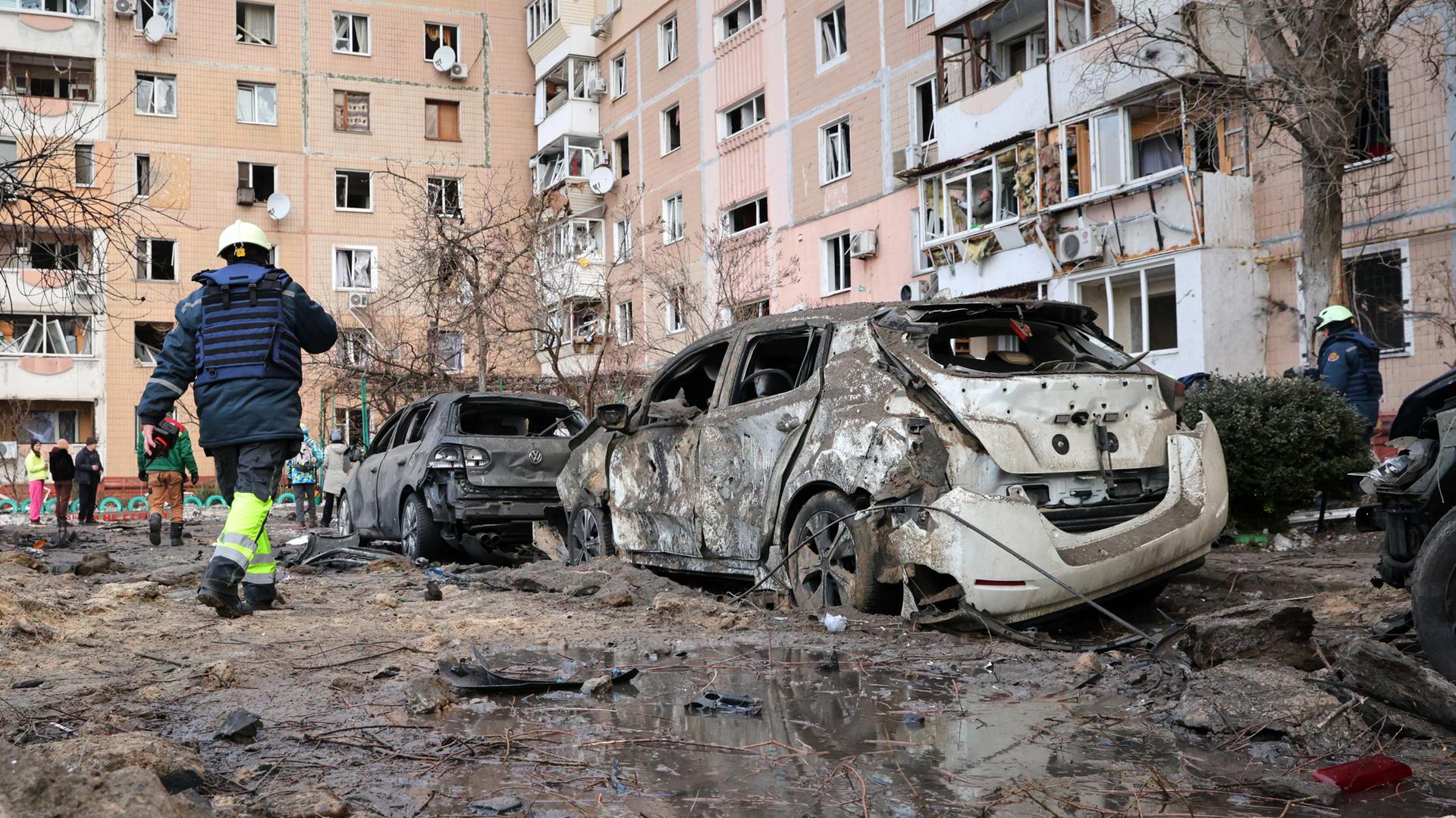 Auf der schlammbedeckten Straße vor einem Wohnblock stehen ausgebrannte Autos. Mehrere Menschen in Gummistiefeln und reflektierender Warnkleidung laufen daran vorbei.