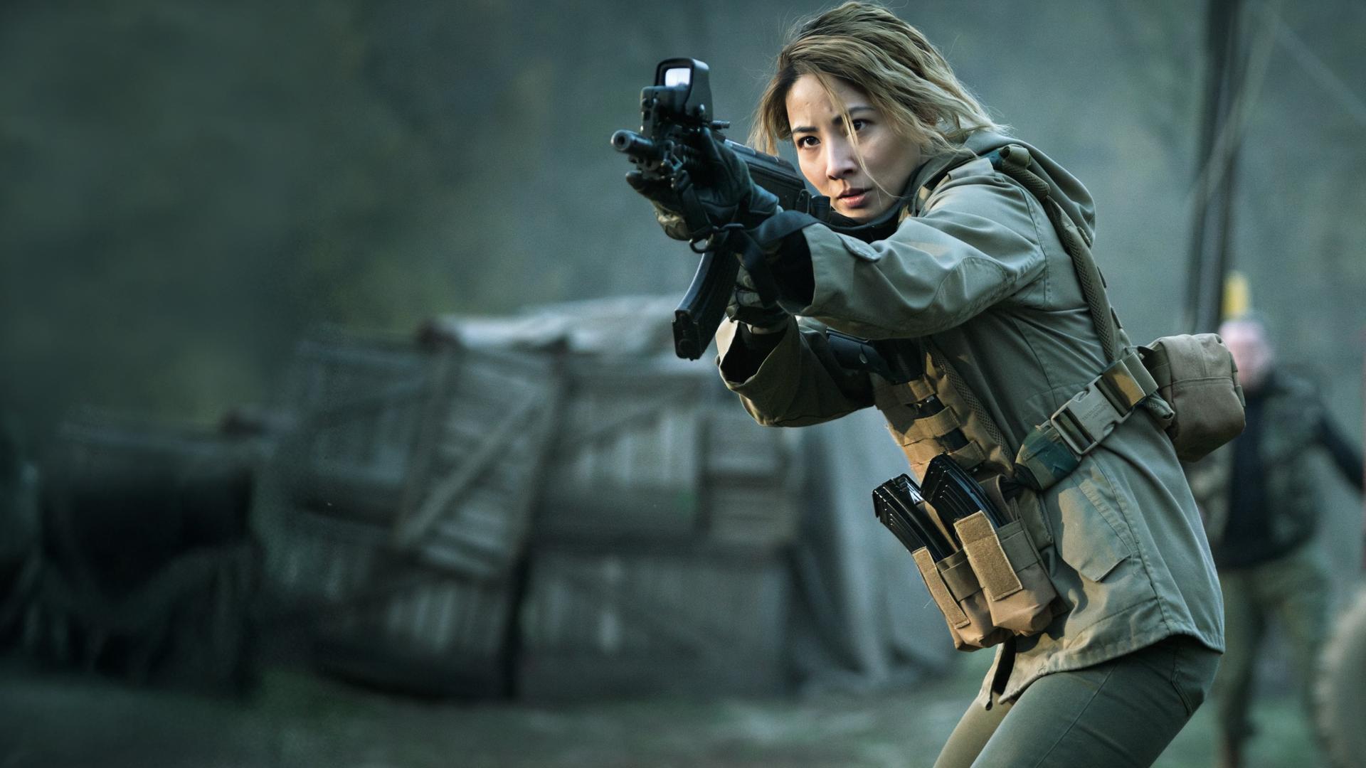 Zada (Jing Lusi) hält ihr Gewehr im Anschlag. Verschwommen im Hintergrund sind ein Traktor, eine weitere Person und abgedeckte Holzkisten zu sehen.