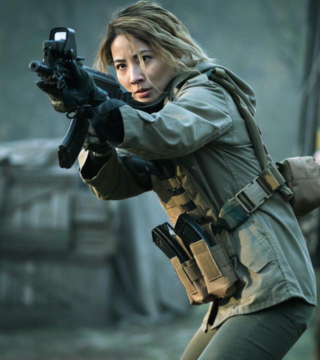 Zada (Jing Lusi) hält ihr Gewehr im Anschlag. Verschwommen im Hintergrund sind ein Traktor, eine weitere Person und abgedeckte Holzkisten zu sehen.