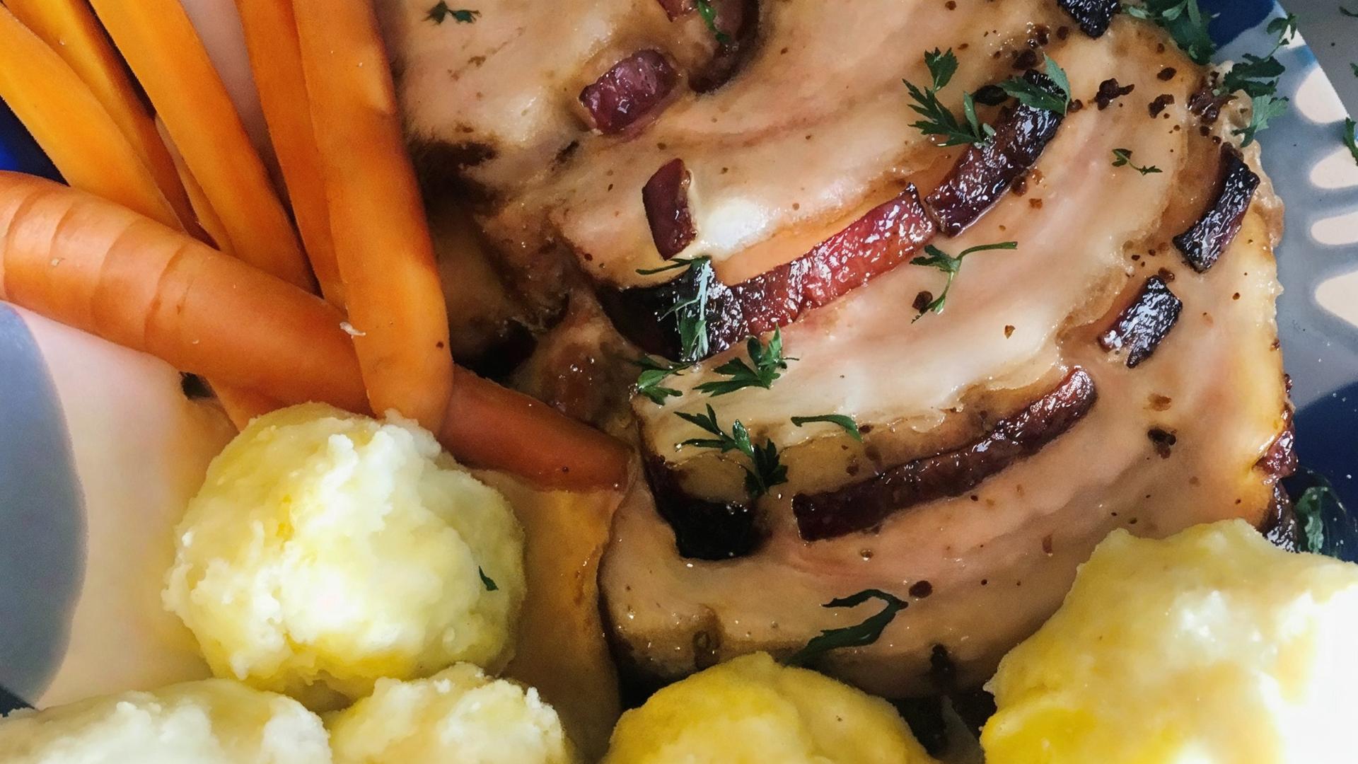 Das Bild zeigt einen Teller mit einem typischen Gericht der österreichischen Küche, insbesondere dem traditionellen "Schweinsbraten". Auf dem Teller liegt eine große, in Scheiben geschnittene Portion Schweinefleisch, das goldbraun und knusprig aussieht. Die Oberfläche des Fleischs ist mit einer dunkleren Kruste versehen und es sind feine Kräuter zur Garnierung sichtbar.  Neben dem Fleisch befinden sich mehrere orangefarbene Karotten, die ungekocht und ungeschält aussehen. Diese sind in länglichen, schmalen Stücken angeordnet.   Auf der anderen Seite des Tellers liegen mehrere knödelähnliche Beilagen, die gelblich und rund sind, wahrscheinlich Mehlknödel. Diese sind weich und haben eine glatte Oberfläche.  Der Teller hat einen farbenfrohen Rand und steht vor einer neutralen Kulisse. Im Hintergrund ist ein kleiner, weißer Schüssel mit frischen Kräutern zu sehen, die als zusätzliche Garnitur dienen könnten.