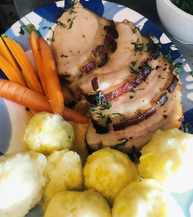 Das Bild zeigt einen Teller mit einem typischen Gericht der österreichischen Küche, insbesondere dem traditionellen "Schweinsbraten". Auf dem Teller liegt eine große, in Scheiben geschnittene Portion Schweinefleisch, das goldbraun und knusprig aussieht. Die Oberfläche des Fleischs ist mit einer dunkleren Kruste versehen und es sind feine Kräuter zur Garnierung sichtbar.  Neben dem Fleisch befinden sich mehrere orangefarbene Karotten, die ungekocht und ungeschält aussehen. Diese sind in länglichen, schmalen Stücken angeordnet.   Auf der anderen Seite des Tellers liegen mehrere knödelähnliche Beilagen, die gelblich und rund sind, wahrscheinlich Mehlknödel. Diese sind weich und haben eine glatte Oberfläche.  Der Teller hat einen farbenfrohen Rand und steht vor einer neutralen Kulisse. Im Hintergrund ist ein kleiner, weißer Schüssel mit frischen Kräutern zu sehen, die als zusätzliche Garnitur dienen könnten.