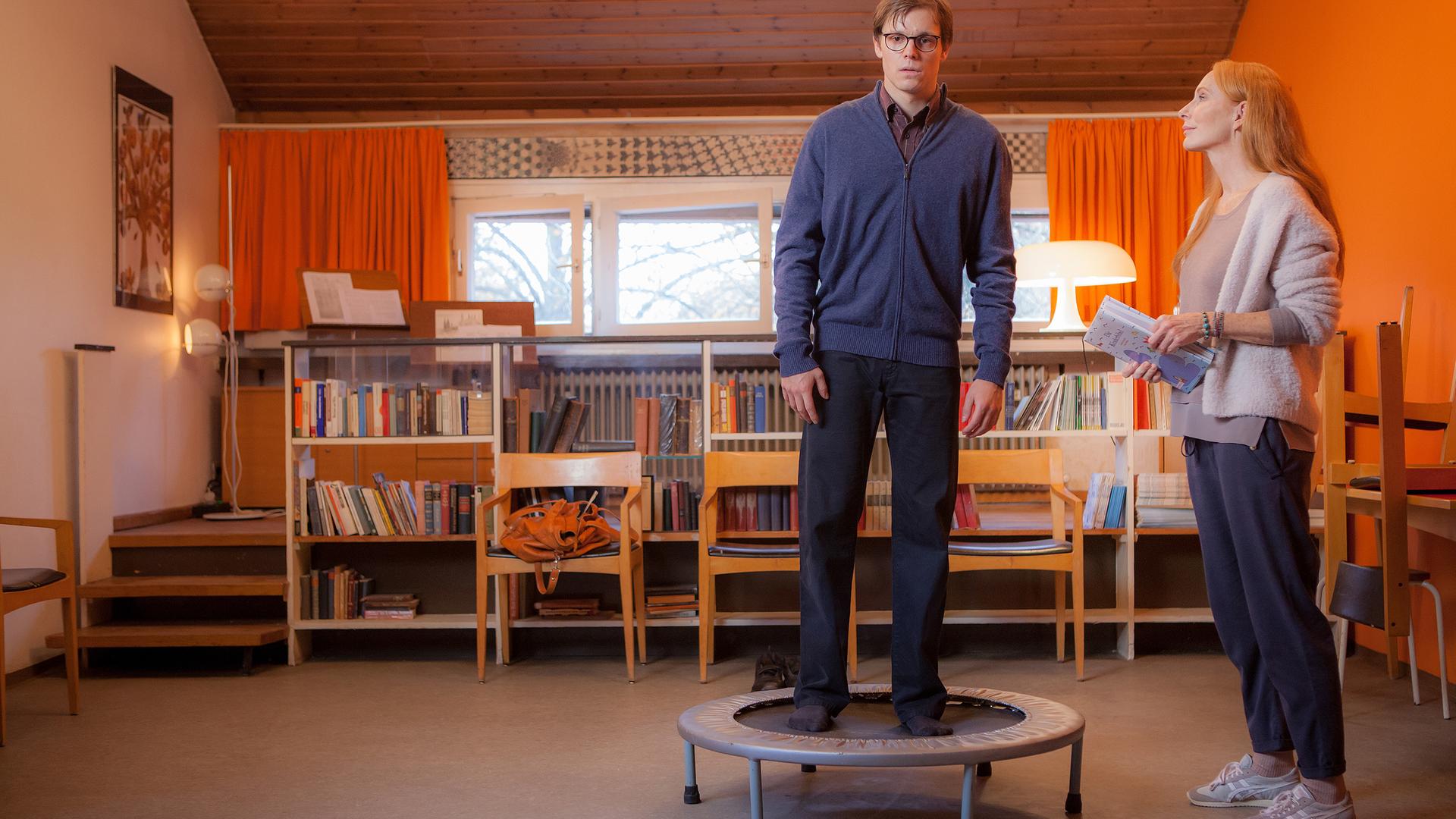 Thaddäus (Franz Hartwig) steht auf dem Trampolin und Rebecca (Andrea Sawatzki) daneben - im Hintergrund Fenster, Bücherregal und Stühle im Seminarraum.