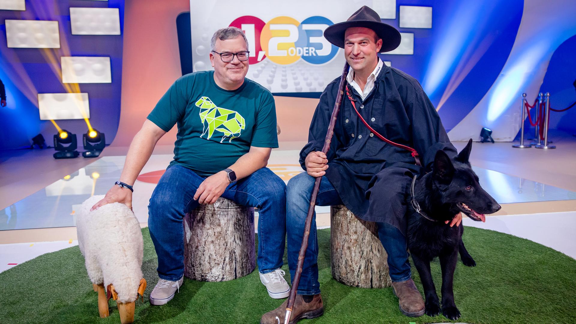 "1, 2 oder 3 - Schafe - määäh-ga spannend": Elton und Wanderschäfer Jonas Henninger sitzen auf Baumstämmen im Studio. Neben Elton stehen zwei Schafe aus Holz. Jonas Henninger streichelt seinem Hund Jackie den Kopf.