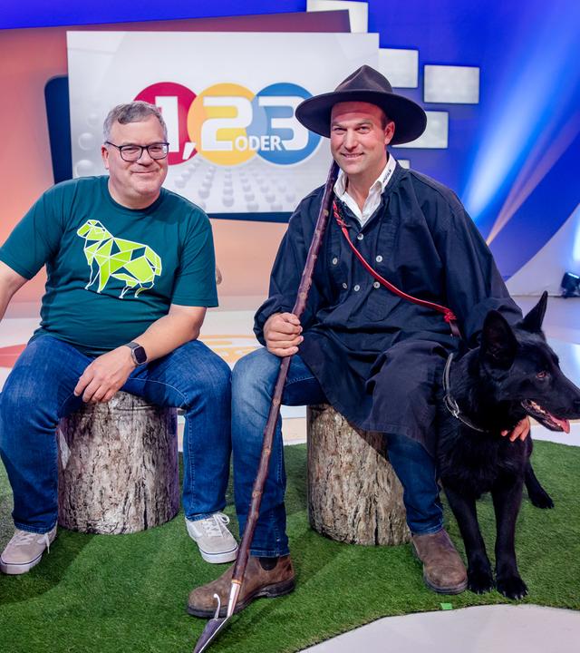 "1, 2 oder 3 - Schafe - määäh-ga spannend": Elton und Wanderschäfer Jonas Henninger sitzen auf Baumstämmen im Studio. Neben Elton stehen zwei Schafe aus Holz. Jonas Henninger streichelt seinem Hund Jackie den Kopf.