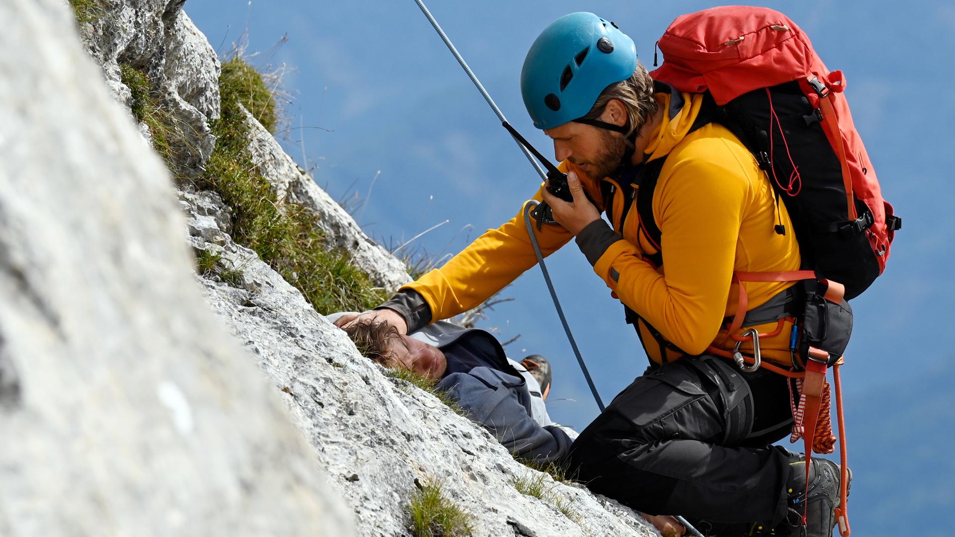 Auf einem Felsvorsprung liegt der verletzte Charly (Julian Waldner). Vor ihm, an einem Seil gesichert, kniet Bergretter Markus (Sebastian Ströbel) und spricht in sein Funkgerät.
