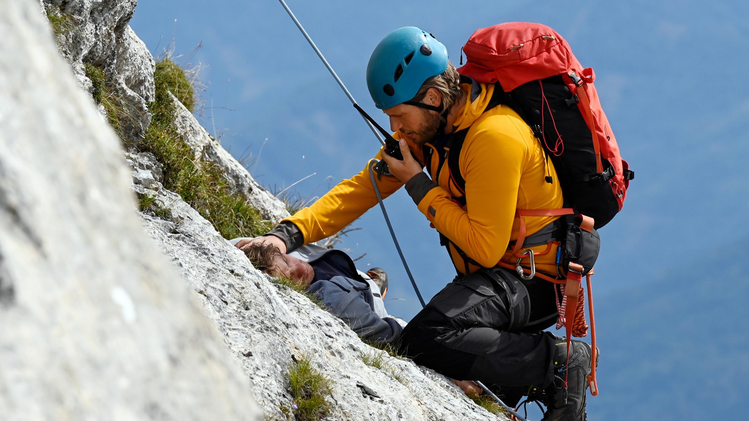 Auf einem Felsvorsprung liegt der verletzte Charly (Julian Waldner). Vor ihm, an einem Seil gesichert, kniet Bergretter Markus (Sebastian Ströbel) und spricht in sein Funkgerät.