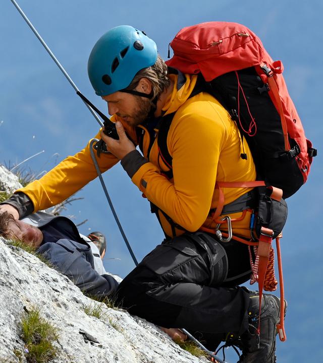 Auf einem Felsvorsprung liegt der verletzte Charly (Julian Waldner). Vor ihm, an einem Seil gesichert, kniet Bergretter Markus (Sebastian Ströbel) und spricht in sein Funkgerät.