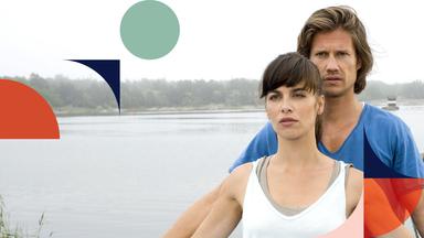 An einem Seeufer - Hanna (Julia-Maria Köhler) steht vor Jonas (Kristian Kiehling). Mit zur Seite ausgestreckten Armen, hält er Hanna an ihren beiden Handgelenken fest. Im Hintergrund ein Bootshaus.