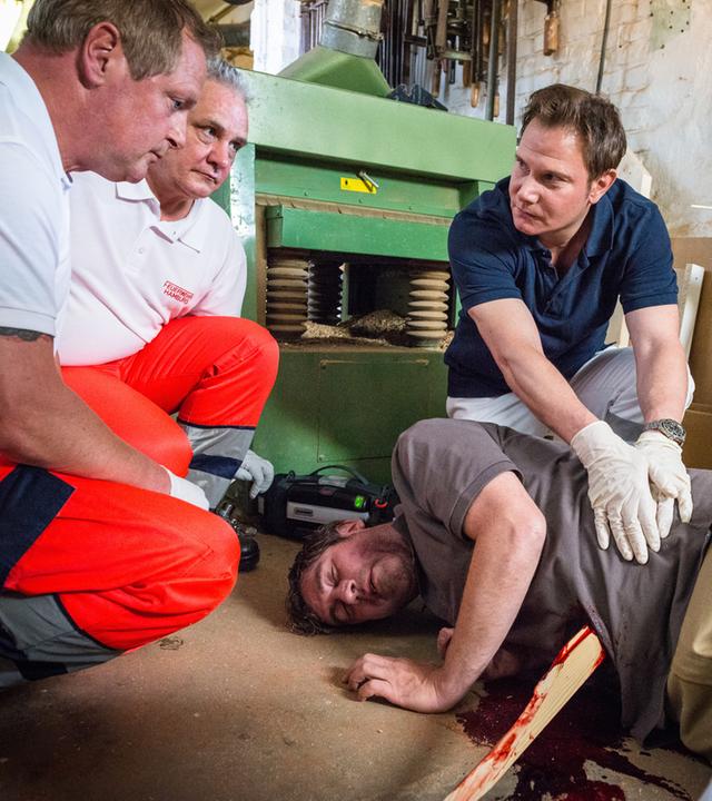 "Notruf Hafenkante - Schatten der Vergangenheit": Auf dem Boden einer Werkstatt liegt ein Verletzter (Stephan Szász) in Seitenlage. Er hat eine Holzleiste im Bauch stecken. Der Notarzt Dr. Haase (Fabian Harloff) kniet hinter ihm und tastet mit beiden Händen den Bauch ab. Seitlich des Verletzten sitzen zwei Rettungssanitäter.