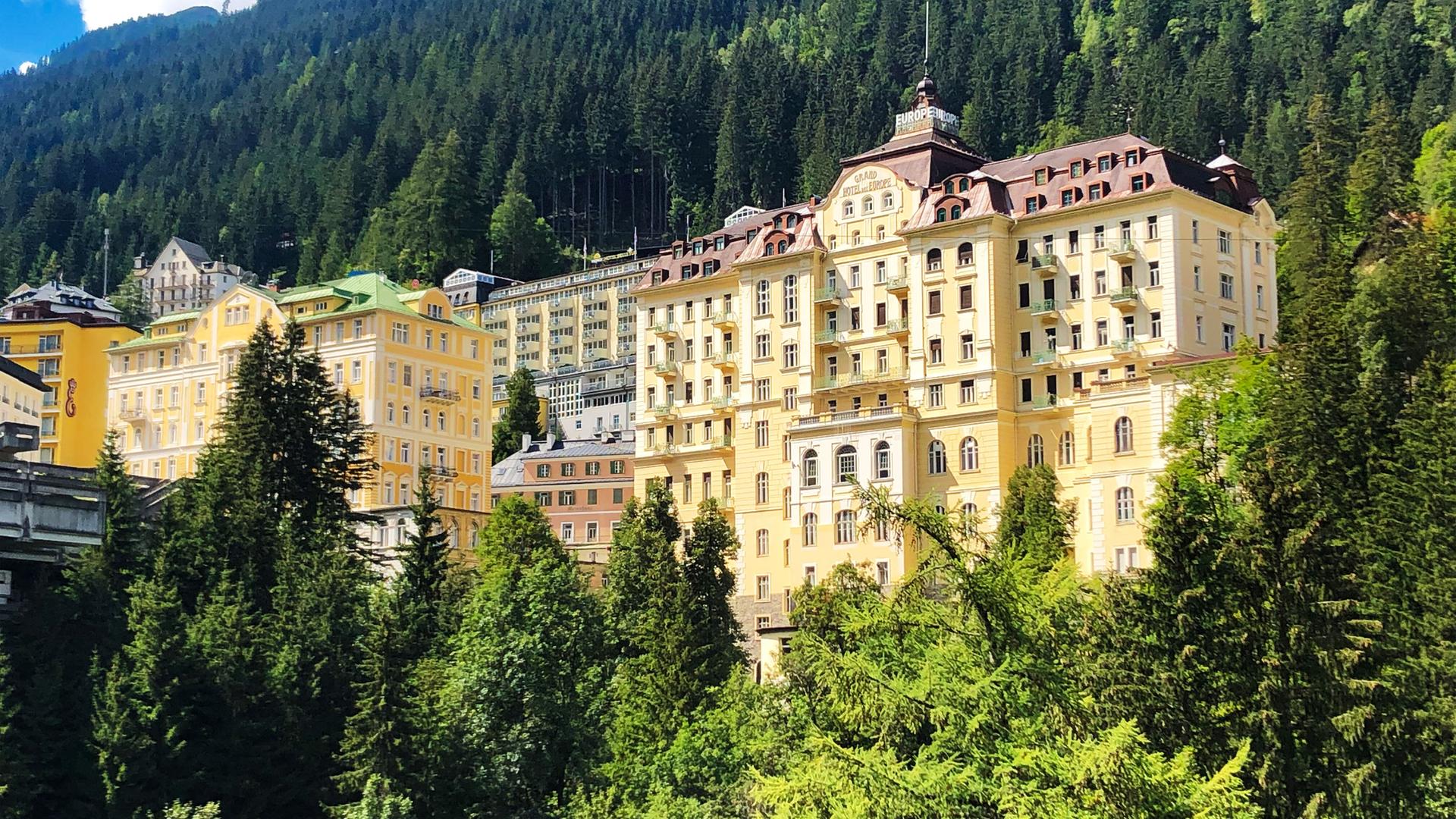 Das Bild zeigt eine Ansicht des Gasteinertals in Österreich, mit einem Fokus auf mehrere große Gebäude, die in einer bergigen Umgebung errichtet sind. Im Vordergrund sind dichte, grüne Bäume sichtbar, während im Hintergrund die bewaldeten Hänge der Berge aufsteigen.   Die Gebäude sind mehrstöckig und haben eine historisch anmutende Architektur, wobei viele von ihnen gelb gestrichen sind. Einige Dächer sind mit Schindeln gedeckt, und es gibt besondere architektonische Details wie Balkone. Das Wetter scheint schön zu sein, mit blauem Himmel und einigen weißen, fluffigen Wolken.  Die Szene vermittelt einen Eindruck von Erholung und Natur, passend zu den Informationen über die heilenden Eigenschaften des Wassers im Gasteinertal, das auch zur Energiegewinnung genutzt wird.