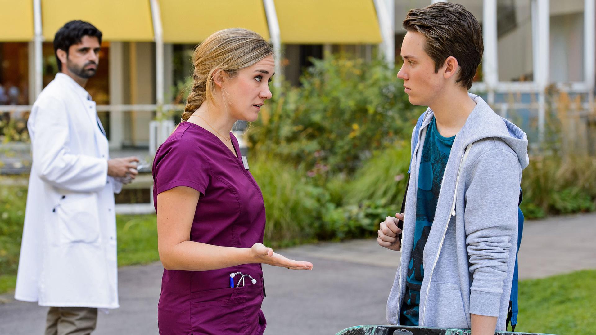 "Bettys Diagnose - Schein und Sein": Betty (Annina Hellenthal) hat mit ihrem Sohn Yannik (Alvar Goetze) vor der Klinik eine Auseinandersetzung. Diese Szene wird von Dr. Behring (Maximilian Grill) im Hintergrund beobachtet.