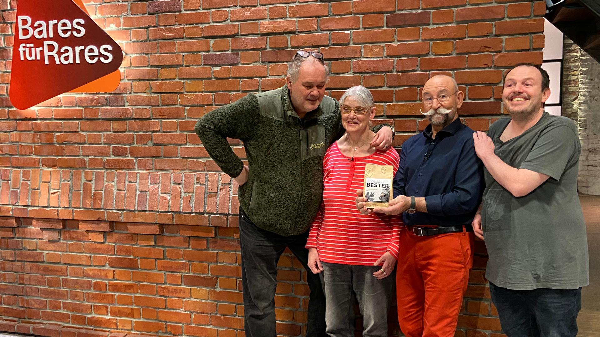 Waldi und Horst Lichter stehen mit zwei Kandidaten von "Bares für Rares" im Studio vor der Backsteinwand. Links ist das "Bares für Rares"-Logo zu sehen.