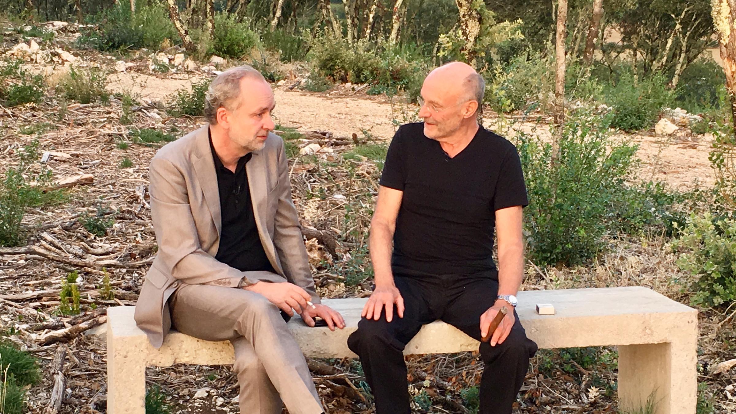 Ferdinand von Schirach und Anselm Kiefer sitzen nebeneinander auf einer Steinbank und unterhalten sich. Beide rauchen.