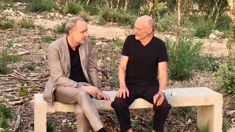 Ferdinand von Schirach und Anselm Kiefer sitzen nebeneinander auf einer Steinbank und unterhalten sich. Beide rauchen.