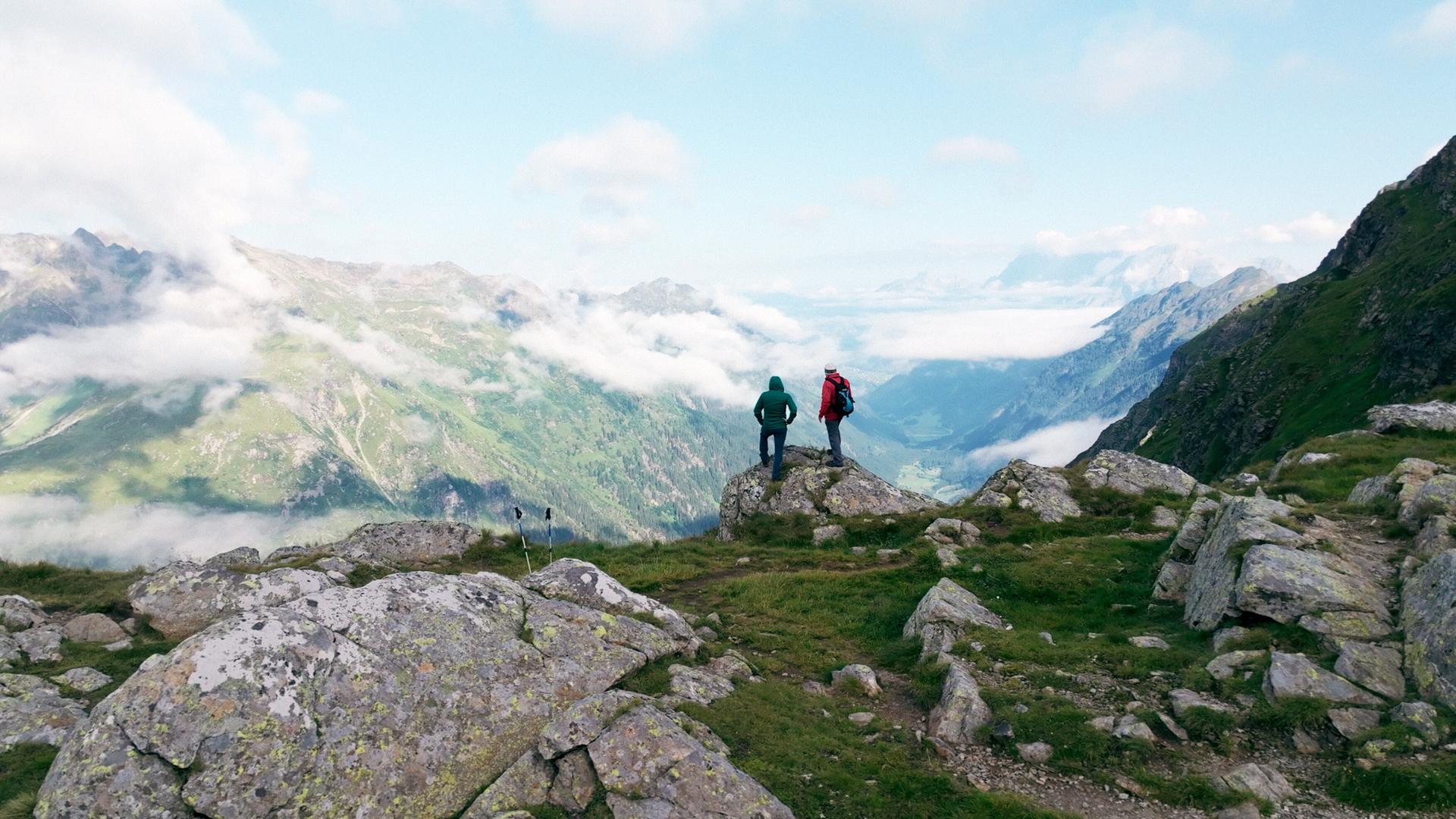 Zwei Wanderer stehen auf einem Felsvorsprung und blicken auf ein weitläufiges, wolkenverhangenes Tal inmitten der Schladminger Tauern; umgeben von felsigem Gelände und grünen Hängen.