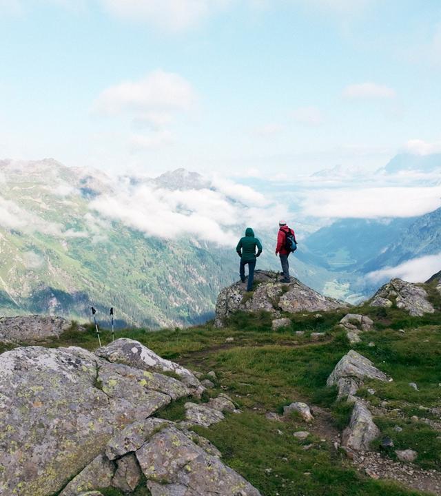 Zwei Wanderer stehen auf einem Felsvorsprung und blicken auf ein weitläufiges, wolkenverhangenes Tal inmitten der Schladminger Tauern; umgeben von felsigem Gelände und grünen Hängen.