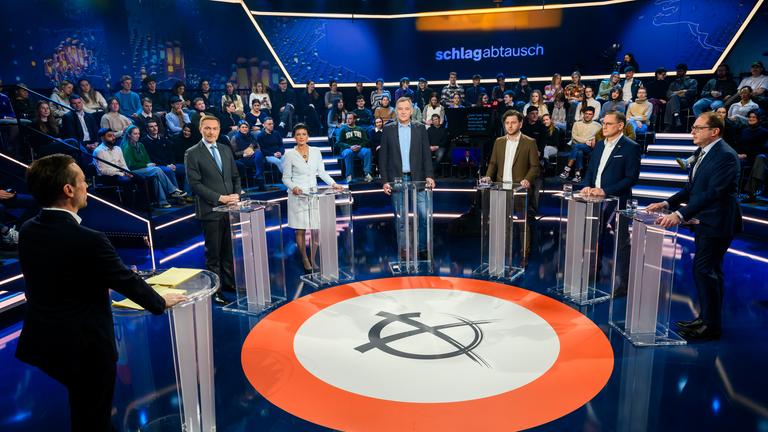 "Schlagabtausch": V.l. Andreas Wunn, Christian Lindner, Sahra Wagenknecht, Jan van Aken, Felix Banaszak, Tino Chrupalla, Alexander Dobrindt stehen im Halbkreis im Studio an Pulten. Im Hintergrund Studiopublikum