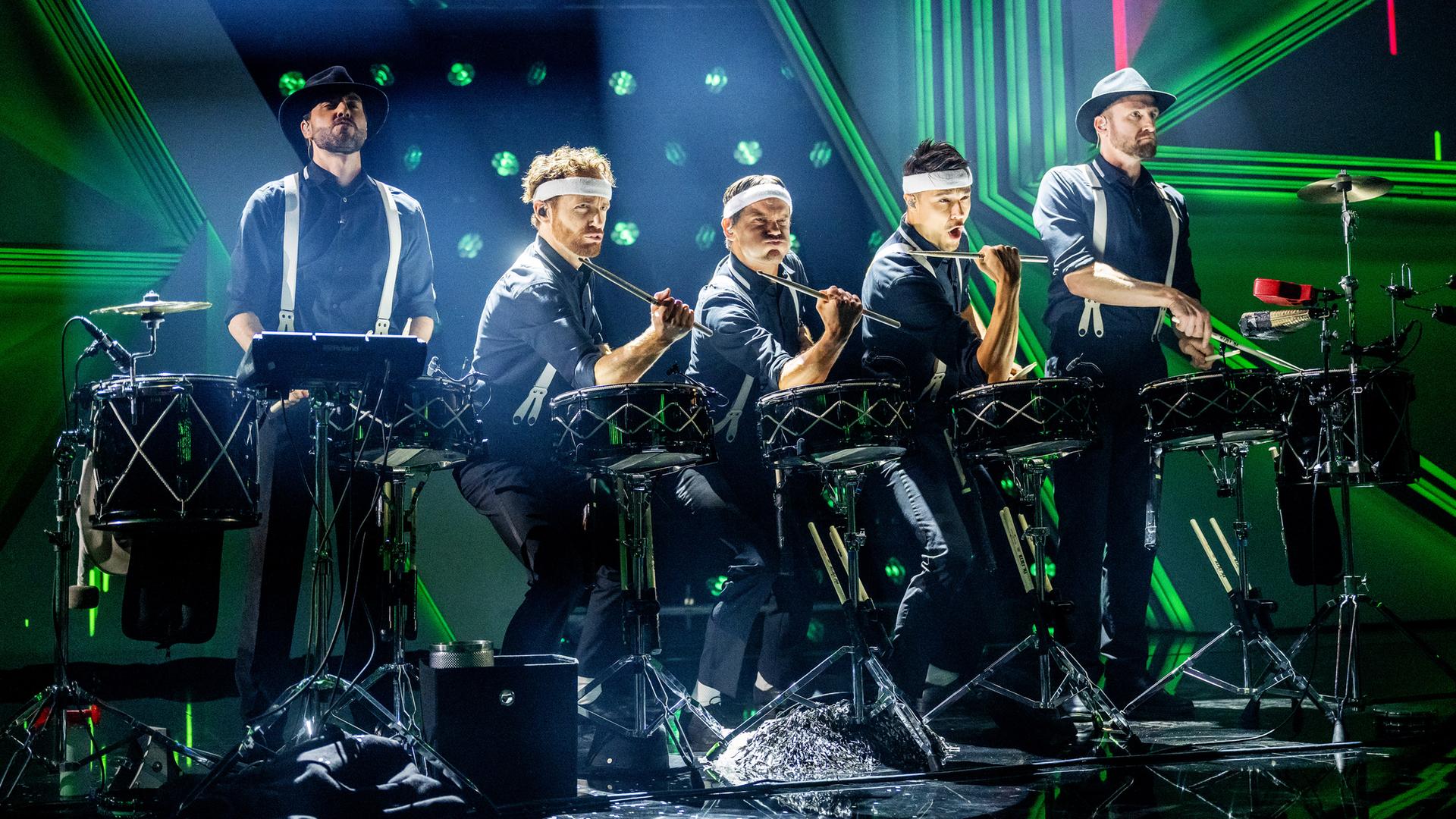 Das Ensemble der Schlagzeugmafia steht in der Mitte der Bühne. Es sind fünf Männer mit Schlagzeugen zu sehen. Der Hintergrund ist blau und grün beleuchtet. 