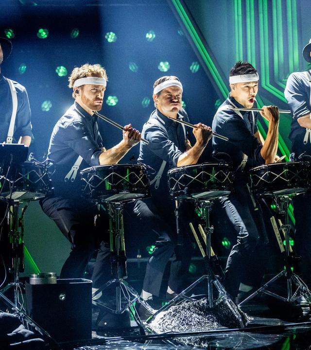 Das Ensemble der Schlagzeugmafia steht in der Mitte der Bühne. Es sind fünf Männer mit Schlagzeugen zu sehen. Der Hintergrund ist blau und grün beleuchtet. 