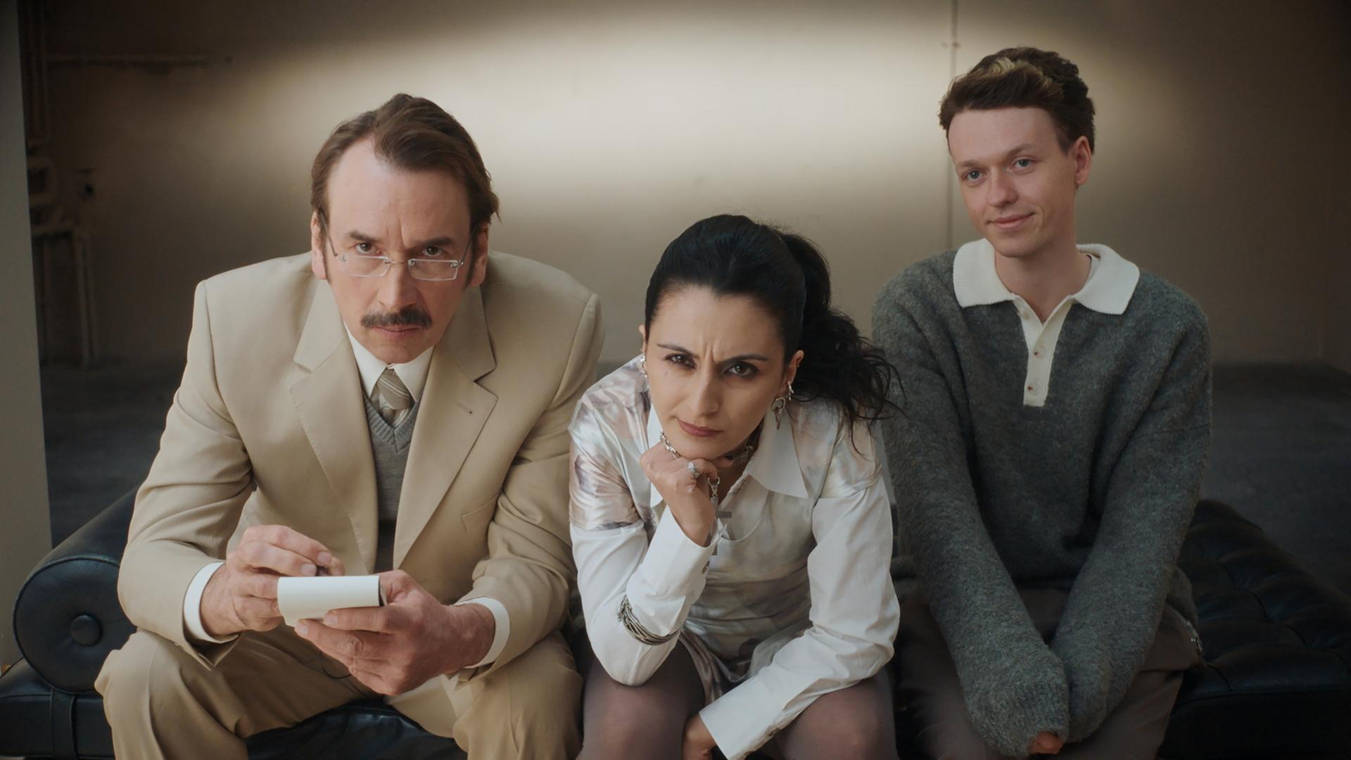 "Schlechte Menschen - Die Perfekte": Cornelius (Arnd Schimkat), Nazlı (Şafak Şengül) und Timoteo (Jeremias Meyer) sitzen nebeneinander auf einer Couch und sehen interessiert in die Kamera.