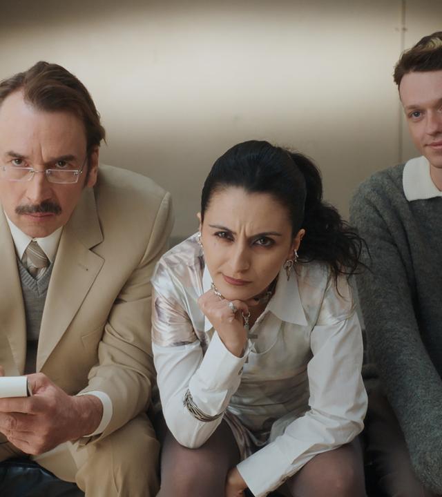 "Schlechte Menschen - Die Perfekte": Cornelius (Arnd Schimkat), Nazlı (Şafak Şengül) und Timoteo (Jeremias Meyer) sitzen nebeneinander auf einer Couch und sehen interessiert in die Kamera.