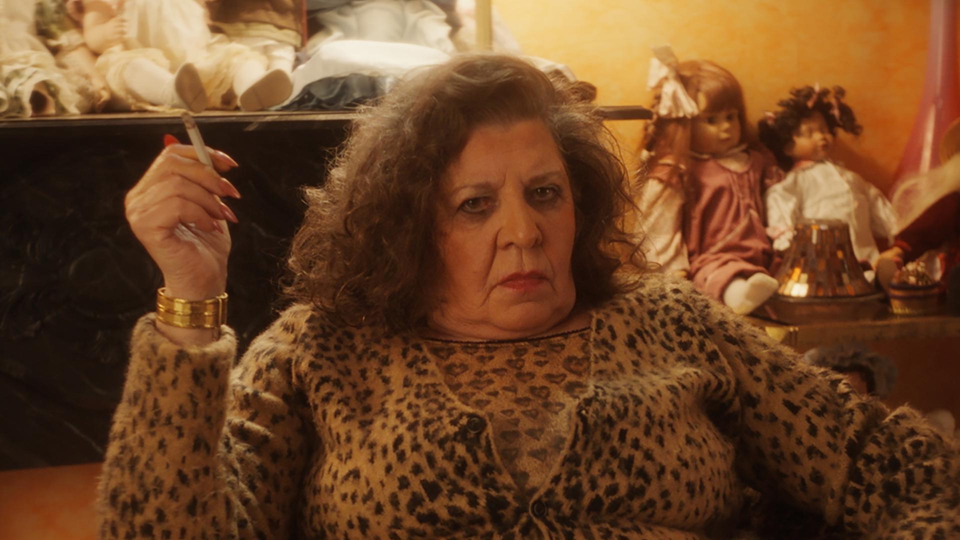 "Schlechte Menschen - Die Mutter": Marie Faruk (Monika Manz) sitzt in Kleidung mit Gepardenlook in einem Sessel in einem Wohnraum und raucht. Hinter ihr sind diverse Regale mit Puppen bevölkert.