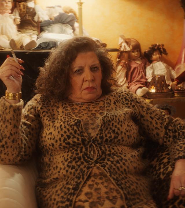 "Schlechte Menschen - Die Mutter": Marie Faruk (Monika Manz) sitzt in Kleidung mit Gepardenlook in einem Sessel in einem Wohnraum und raucht. Hinter ihr sind diverse Regale mit Puppen bevölkert.