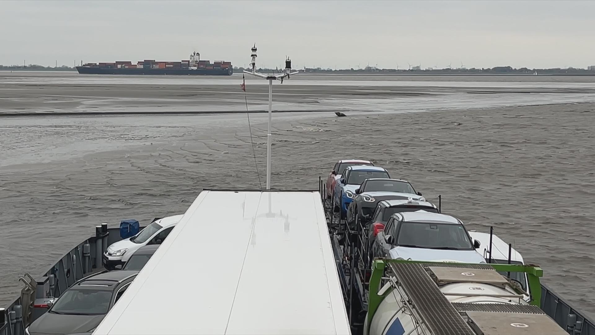 Teil einer Elbfähre, die im Schlick feststeckt. Im Hintergrund ist ein großes Conteinerschiff zu sehen. In der Elbe herrscht Ebbe.