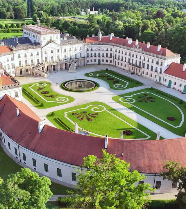 Das Bild zeigt eine Luftaufnahme des Schlosses Esterházy in Fertőd, Ungarn. Im Vordergrund ist ein weitläufiger Schlosspark mit geometrisch angelegten Gärten zu sehen, die mit grünen, runden Hecken und bunten Blumenbeeten geschmückt sind. Das Schloss selbst besteht aus mehreren Gebäudeteilen, die eine elegante, blasse Fassade aufweisen, gekennzeichnet durch große Fenster und mit einem Dach aus roten Ziegeln. In der Mitte des Innenhofs befindet sich ein runder Brunnen, umgeben von geschwungenen Gartenwegen. Im Hintergrund erstreckt sich eine grüne Waldlandschaft. Die Architektur und die Gartenanlagen verleihen dem Ort eine majestätische, barocke Atmosphäre.