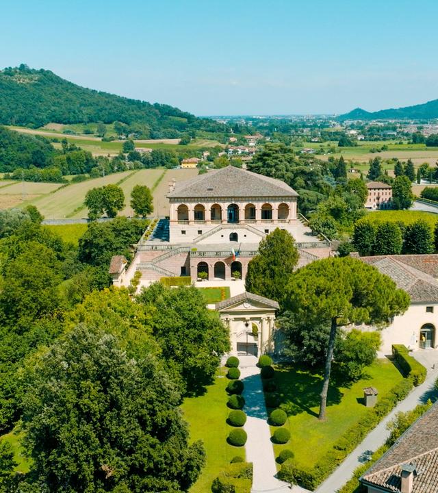 Die Villa Vescovi liegt eingebettet in die Weinlandschaft der Euganeischen Hügel. Das historische Gebäude mit seiner markanten Loggia thront inmitten gepflegter Gartenanlagen, umgeben von sanften Hügeln und grüner Vegetation.