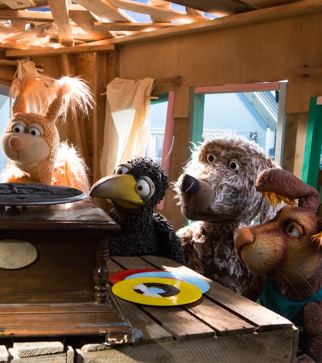 Wurm Lotti, mit Sturzhelm auf dem Kopf, liegt auf einem Plattenteller im Baumhaus. Um sie versammelt sind die Freunde der Rasselbande, sie schauen gebannt auf Lotti. 