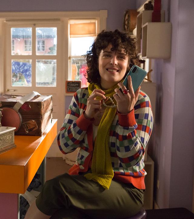 Rabe Rudi sitzt im Laden auf der Kasse und schaut zu Frau Siebenstein, die in ihr Handy schaut.