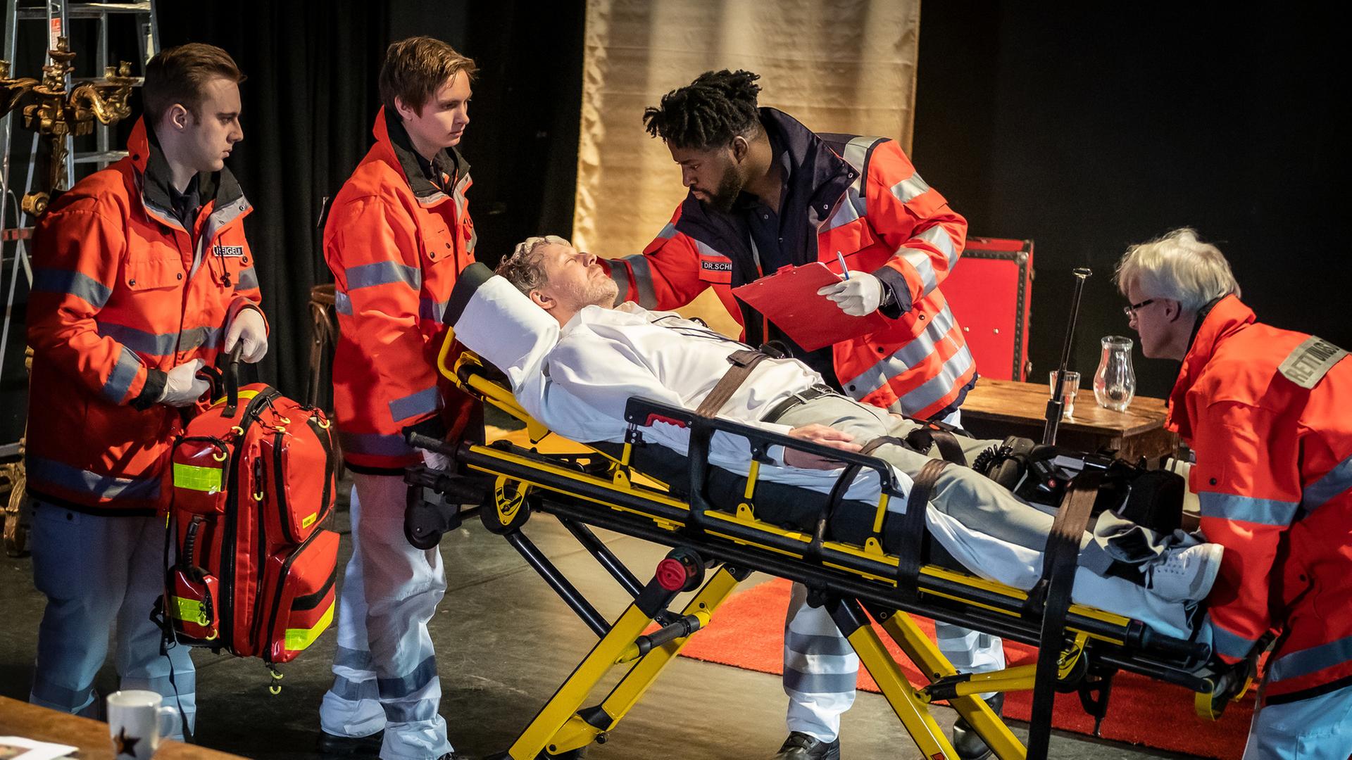 "Notruf Hafenkante - Schluss mit lustig!": Daniel Lohse (Jörn Knebel) liegt auf einer Patiententrage. Dr. Schmidt (Joel Williams) steht neben ihm. Er hat sich über den Patienten gebeugt, seine rechte Hand liegt auf seinem Kopf. In der Linken hält er ein Klemmbrett und einen Stift. Mehrere Rettungssanitäter (Komparsen) sind im Einsatz.
