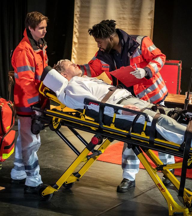 "Notruf Hafenkante - Schluss mit lustig!": Daniel Lohse (Jörn Knebel) liegt auf einer Patiententrage. Dr. Schmidt (Joel Williams) steht neben ihm. Er hat sich über den Patienten gebeugt, seine rechte Hand liegt auf seinem Kopf. In der Linken hält er ein Klemmbrett und einen Stift. Mehrere Rettungssanitäter (Komparsen) sind im Einsatz.