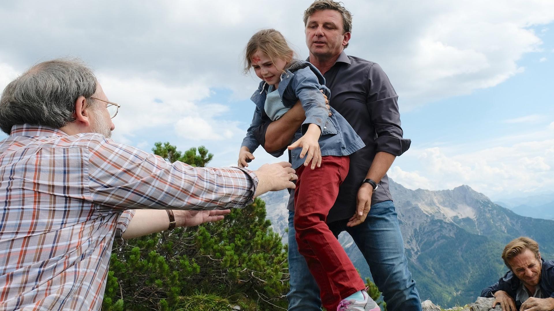 An einer Felskante hält sich Lennart (Gabriel Raab) mit aller Kraft fest. Dr. Martin Gruber (Hans Sigl) trägt die kleine Greta (Amelie Klein), die an der Stirn blutet, zu ihrem Großvater Ulrich (Rainer Bock), der ihr die Arme entgegen streckt.
