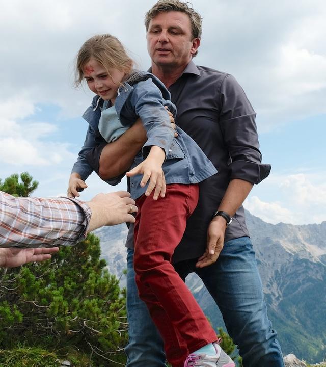 An einer Felskante hält sich Lennart (Gabriel Raab) mit aller Kraft fest. Dr. Martin Gruber (Hans Sigl) trägt die kleine Greta (Amelie Klein), die an der Stirn blutet, zu ihrem Großvater Ulrich (Rainer Bock), der ihr die Arme entgegen streckt.