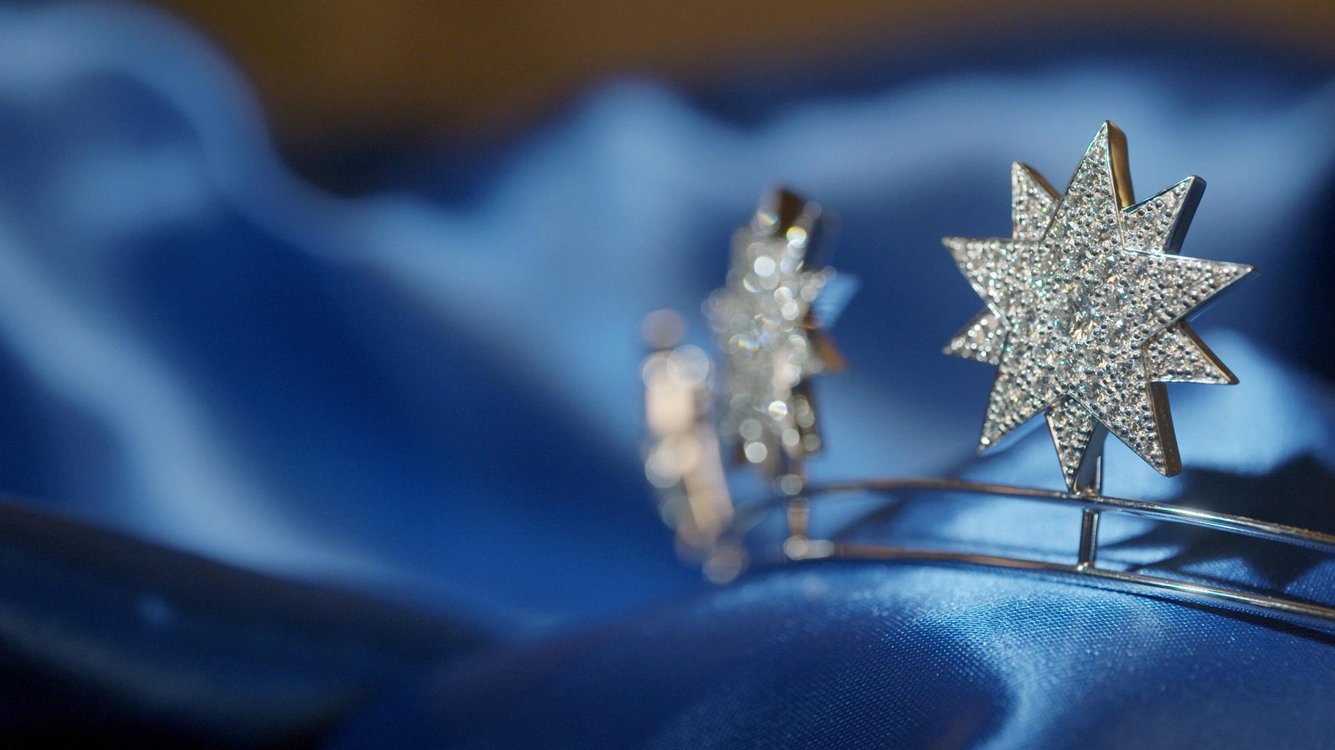 Das Bild zeigt eine Nahaufnahme von zwei funkelnden, sternförmigen Schmuckstücken, die in einem glänzenden blauen Stoff gebettet sind. Die Sterne bestehen aus dem glänzenden Material, das mit vielen kleinen, glitzernden Steinen besetzt ist, was ihnen einen eleganten und opulenten Eindruck verleiht. Der Hintergrund ist weich verschwommen und hebt die Schmuckstücke deutlich hervor. Die Gesamtkomposition vermittelt ein Gefühl von Luxus und Raffinesse, passend zum Thema des Hofjuweliers und der Pracht der Monarchen.