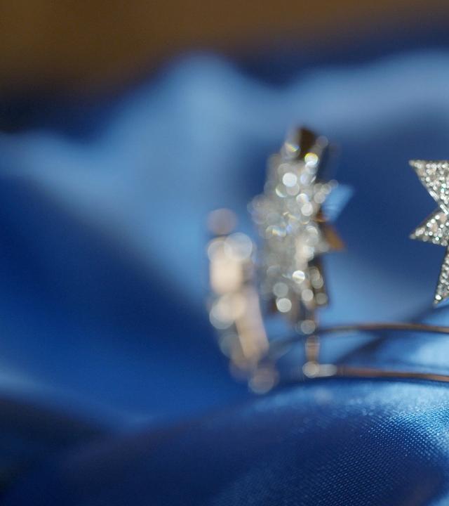 Das Bild zeigt eine Nahaufnahme von zwei funkelnden, sternförmigen Schmuckstücken, die in einem glänzenden blauen Stoff gebettet sind. Die Sterne bestehen aus dem glänzenden Material, das mit vielen kleinen, glitzernden Steinen besetzt ist, was ihnen einen eleganten und opulenten Eindruck verleiht. Der Hintergrund ist weich verschwommen und hebt die Schmuckstücke deutlich hervor. Die Gesamtkomposition vermittelt ein Gefühl von Luxus und Raffinesse, passend zum Thema des Hofjuweliers und der Pracht der Monarchen.